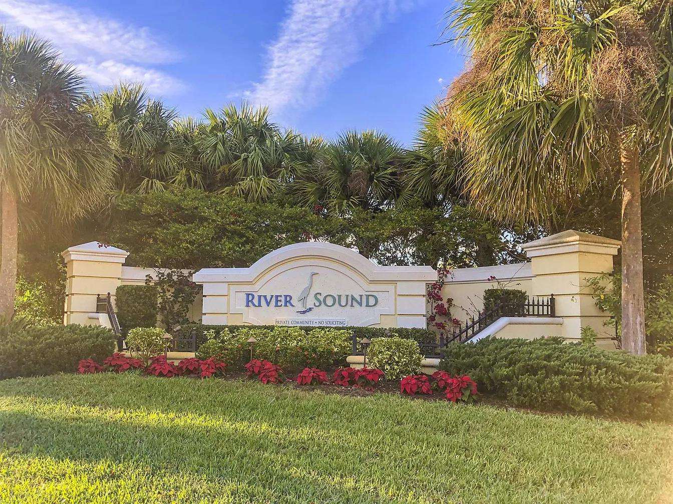 Landscaped gated community entrance with palm trees, green shrubs, and a cream stone sign reading "River Sound." - property