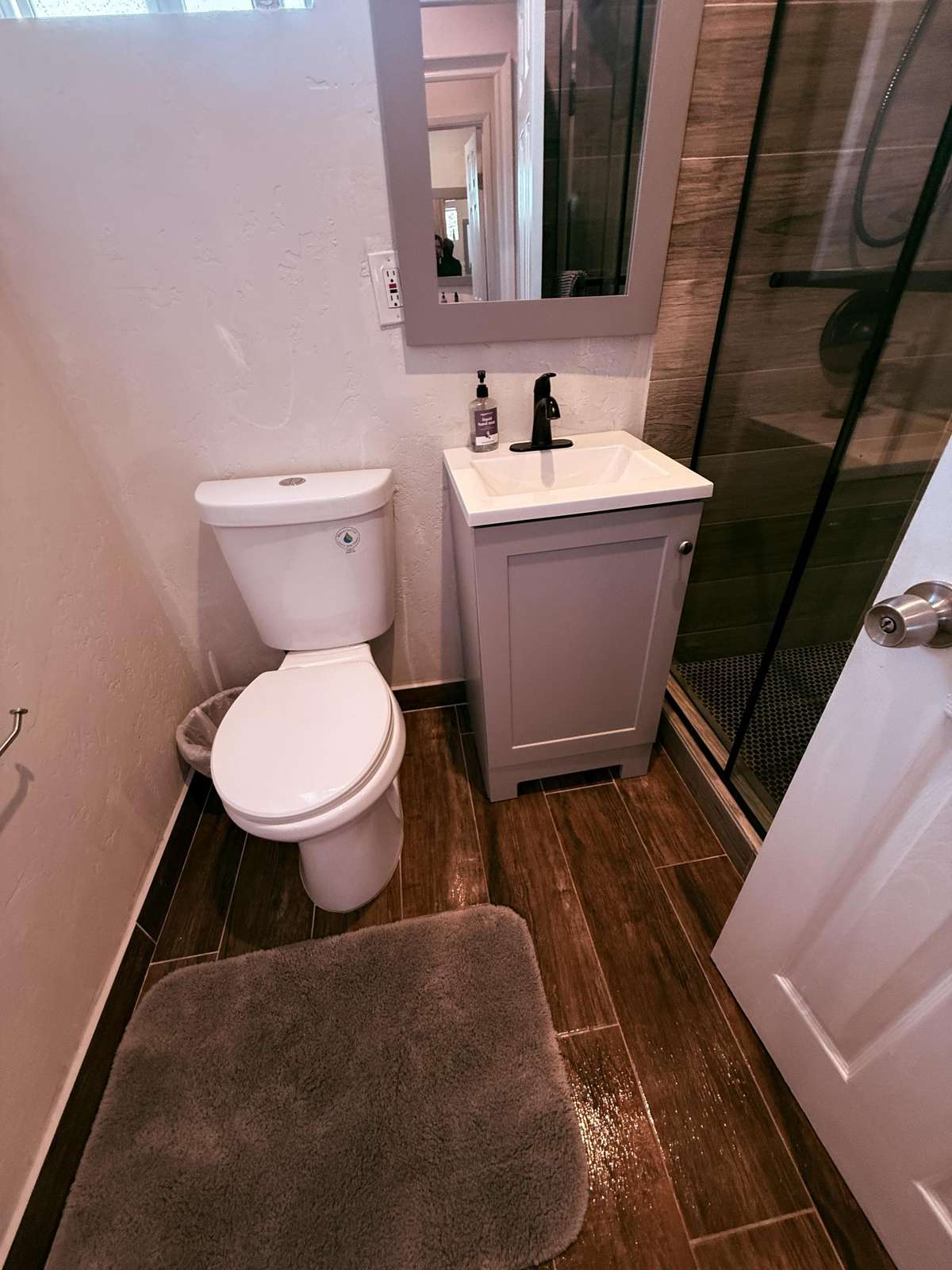 Bathroom with wood-look tile flooring, gray vanity under a framed mirror, and glass-enclosed shower with dark fixtures.