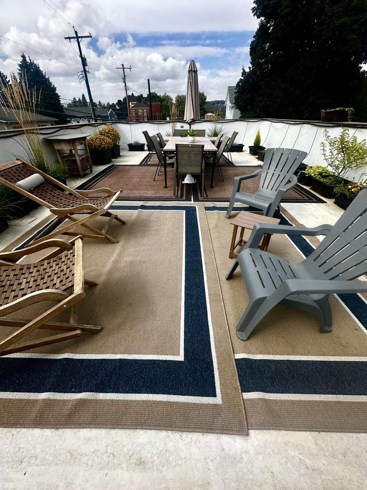 Outdoor rooftop deck featuring a dining table with umbrella, woven lounge chairs, Adirondack seats, and potted plants.