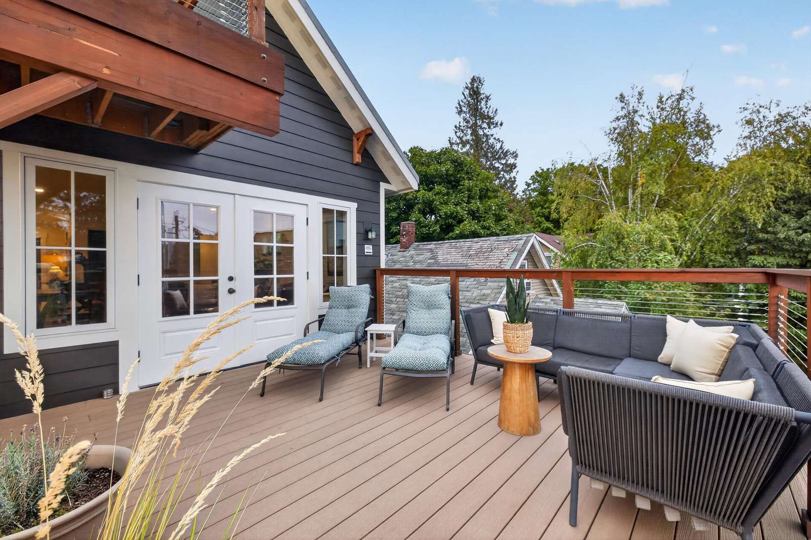 Upper-level deck with gray sectional sofa, black rope chairs, blue cushioned loungers, and cable railing.