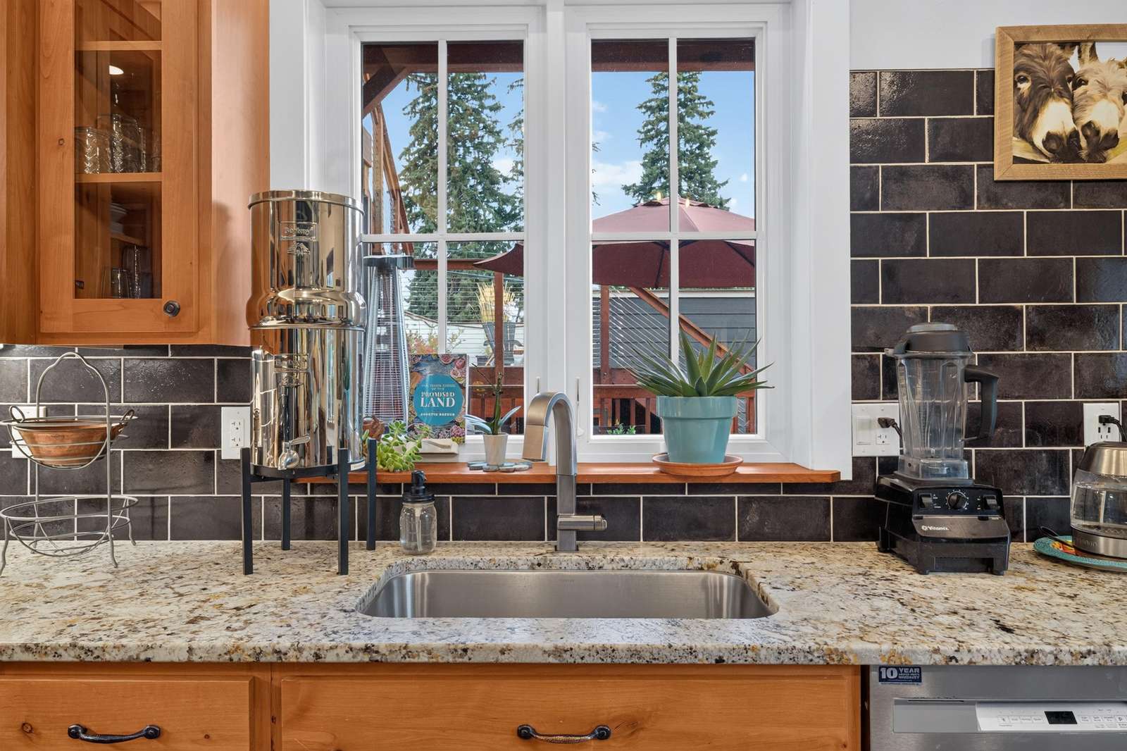 Granite countertop kitchen sink framed by black subway tile backsplash and wood cabinets beneath a white window.