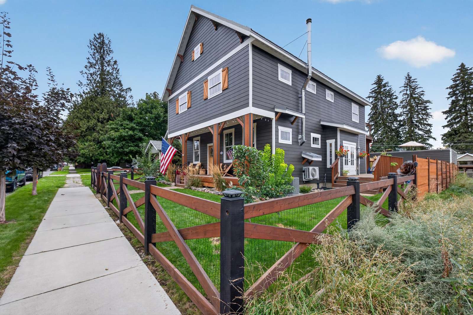 Three-story gray house with white trim, wood shutters, and a fenced green lawn along a concrete sidewalk.