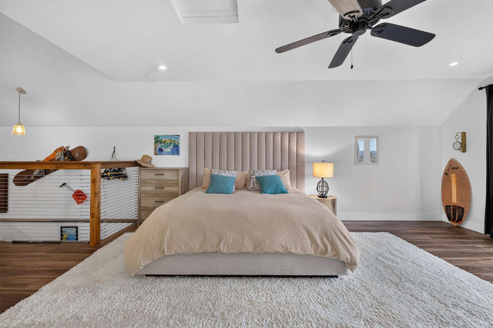 Loft bedroom with tall beige headboard, soft bedding, ceiling fan, and skateboards displayed on wood railing.