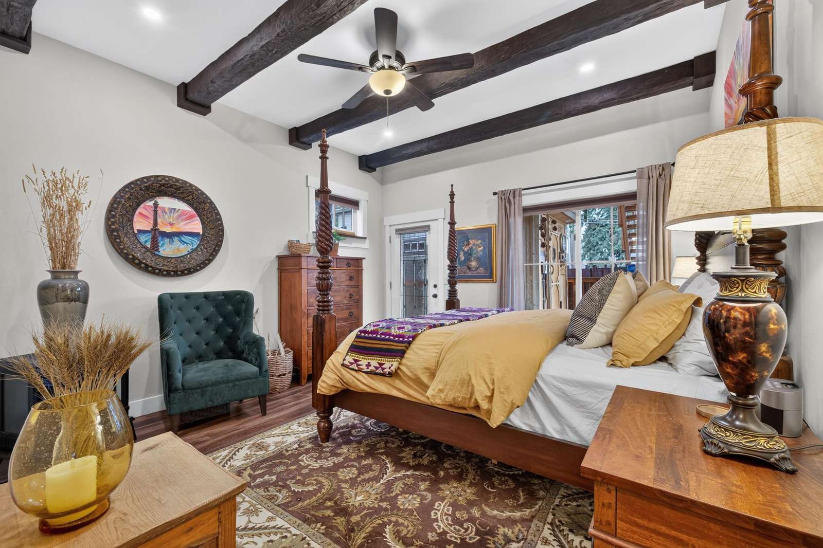 Four-poster bed with mustard bedding sits on patterned rug beneath dark wood ceiling beams and ceiling fan.