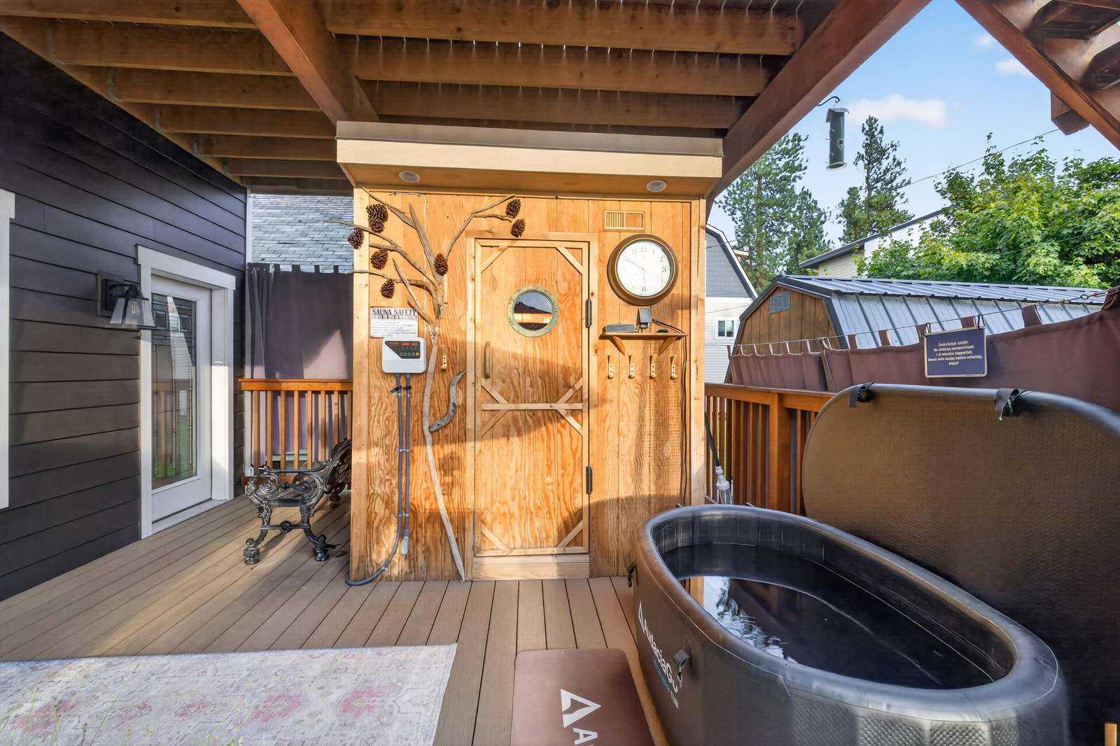 Covered deck with inflatable hot tub, wooden sauna door, pinecone wall art, and black metal bench seating.