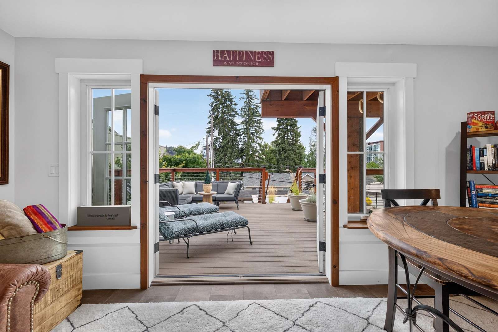Open double doors lead to a spacious deck with cushioned chaise lounges, sectional sofa, and potted plants.