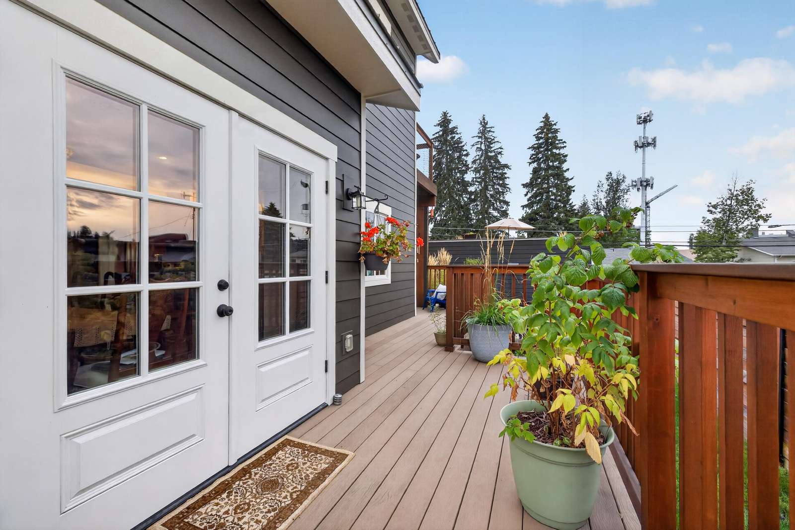 Deck area with wood railing, potted plants, hanging flowers, and white French doors reflecting sunset sky.