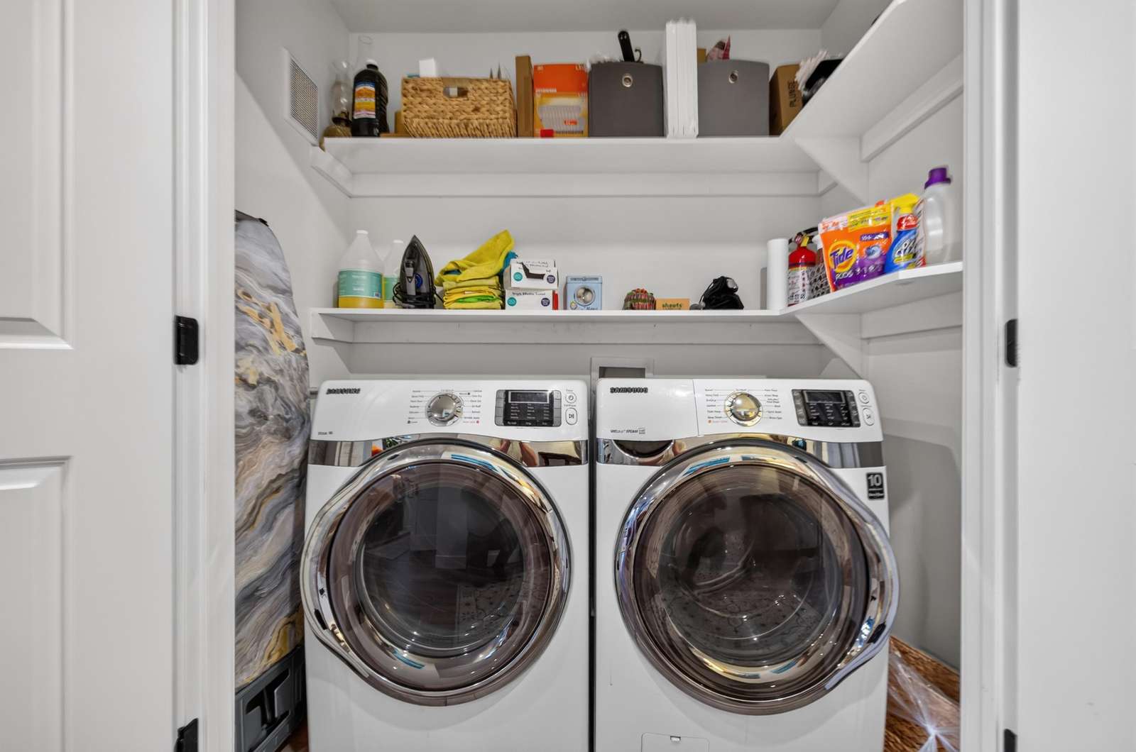 Laundry area with front-loading washer and dryer beneath white shelves stocked with cleaning supplies and linens.