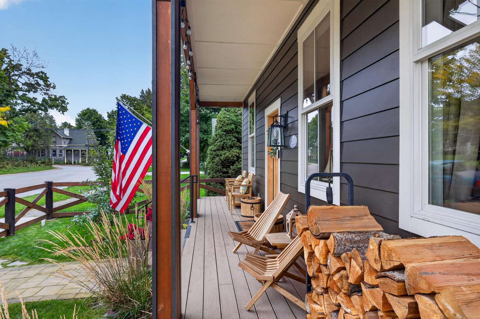 Long front porch with wood Adirondack chairs, stacked firewood, and an American flag against dark siding.