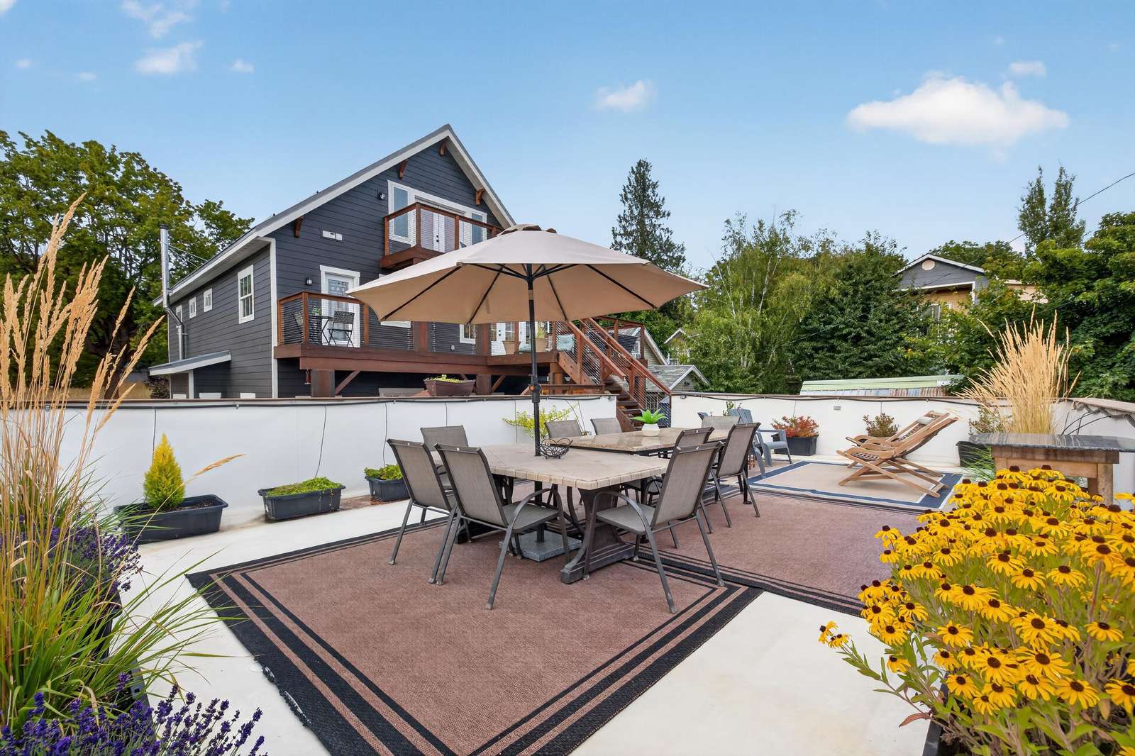Concrete patio with outdoor dining table, umbrella, lounge chairs, and planters with yellow flowers.