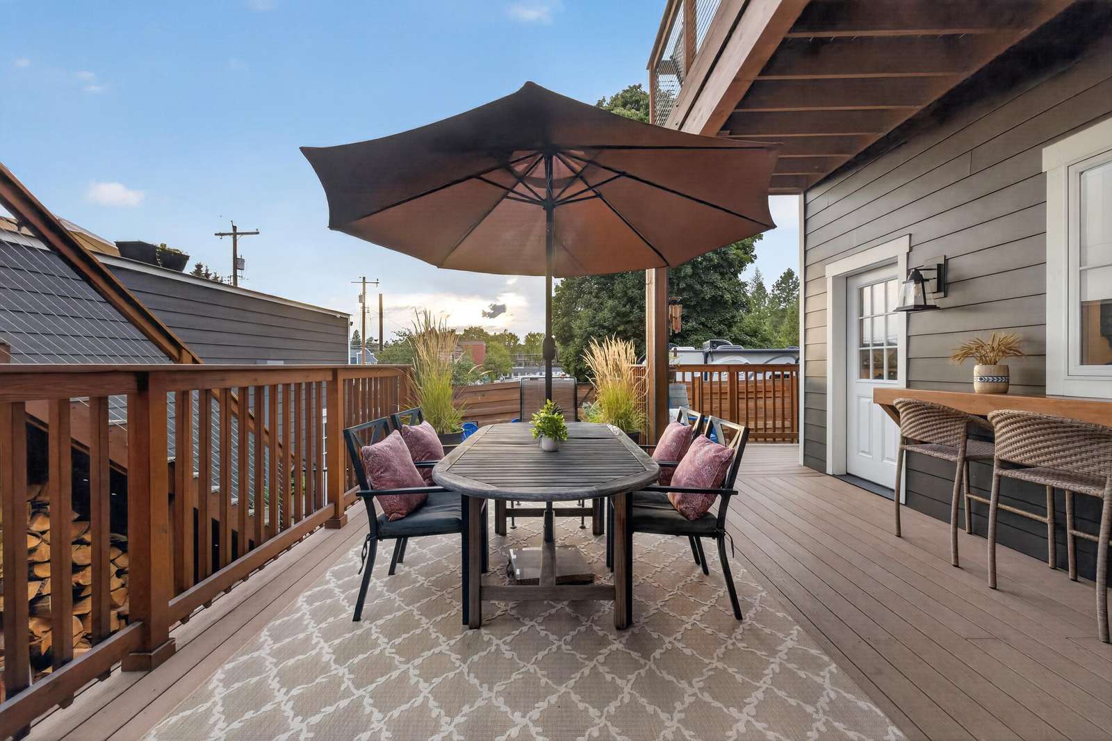 Deck dining area with dark wood table, cushioned chairs, large umbrella, and woven bar stools along siding wall.
