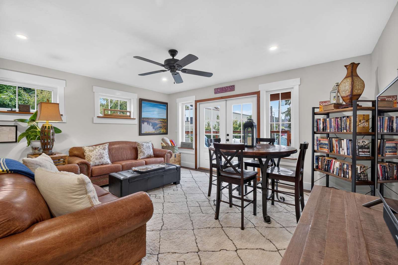 Living room with brown leather sofas, patterned area rug, dark wood dining table, and bookshelves under recessed lights.