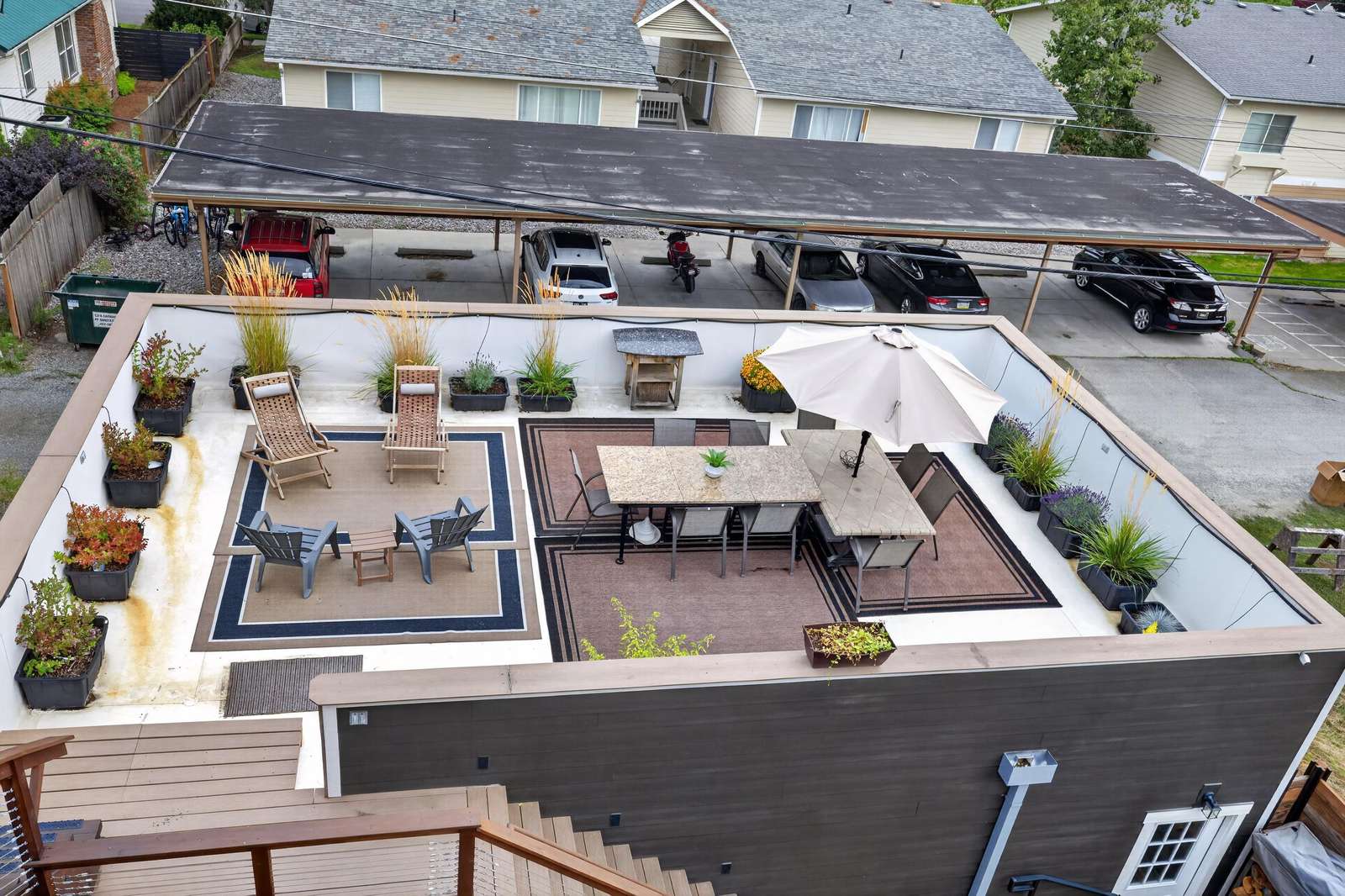 Rooftop patio with two woven lounge chairs, four gray Adirondacks, dining table, umbrella, and potted plants.