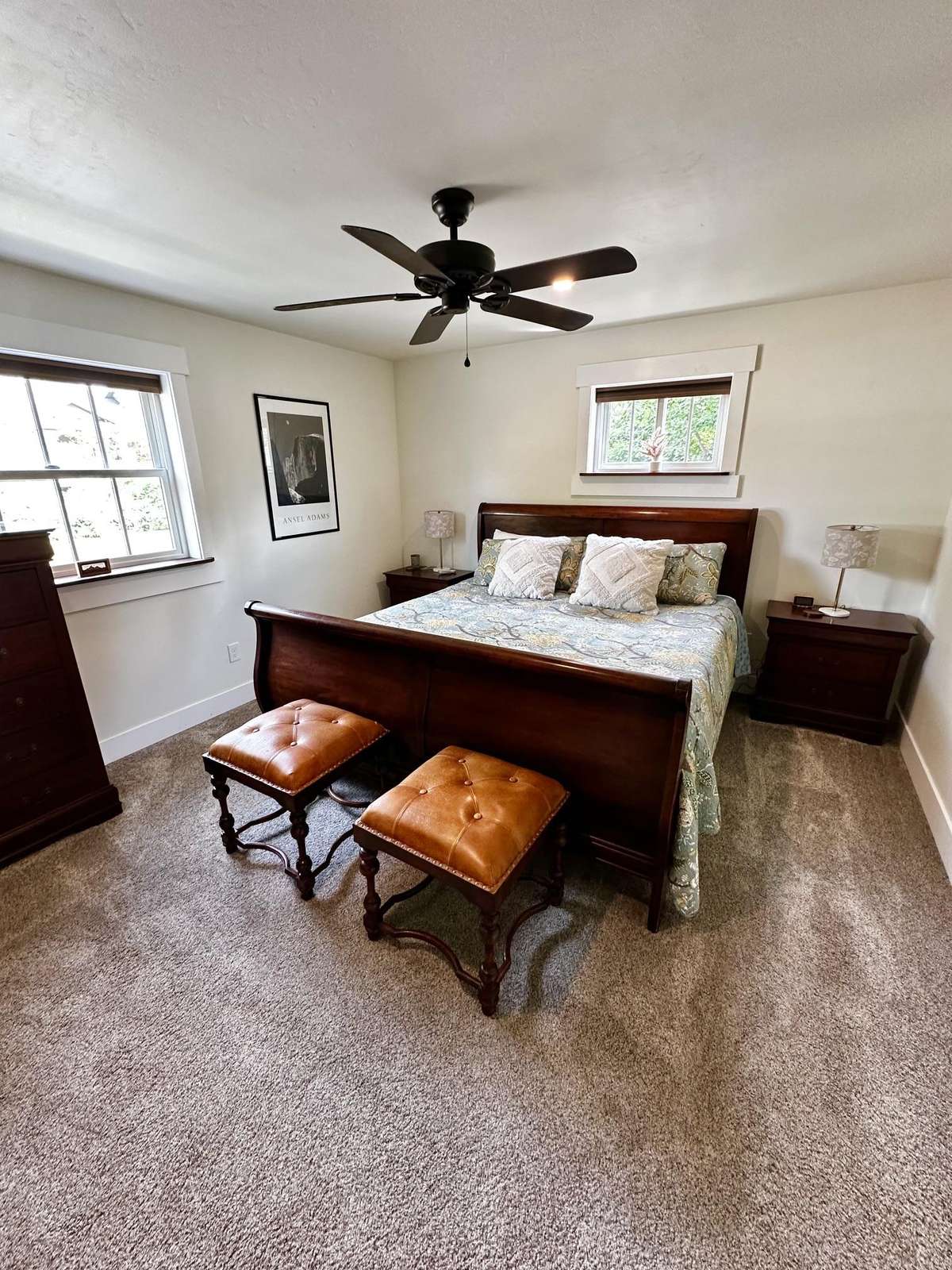 Master bedroom with dark wood sleigh bed, tufted leather stools, and neutral carpet flooring.