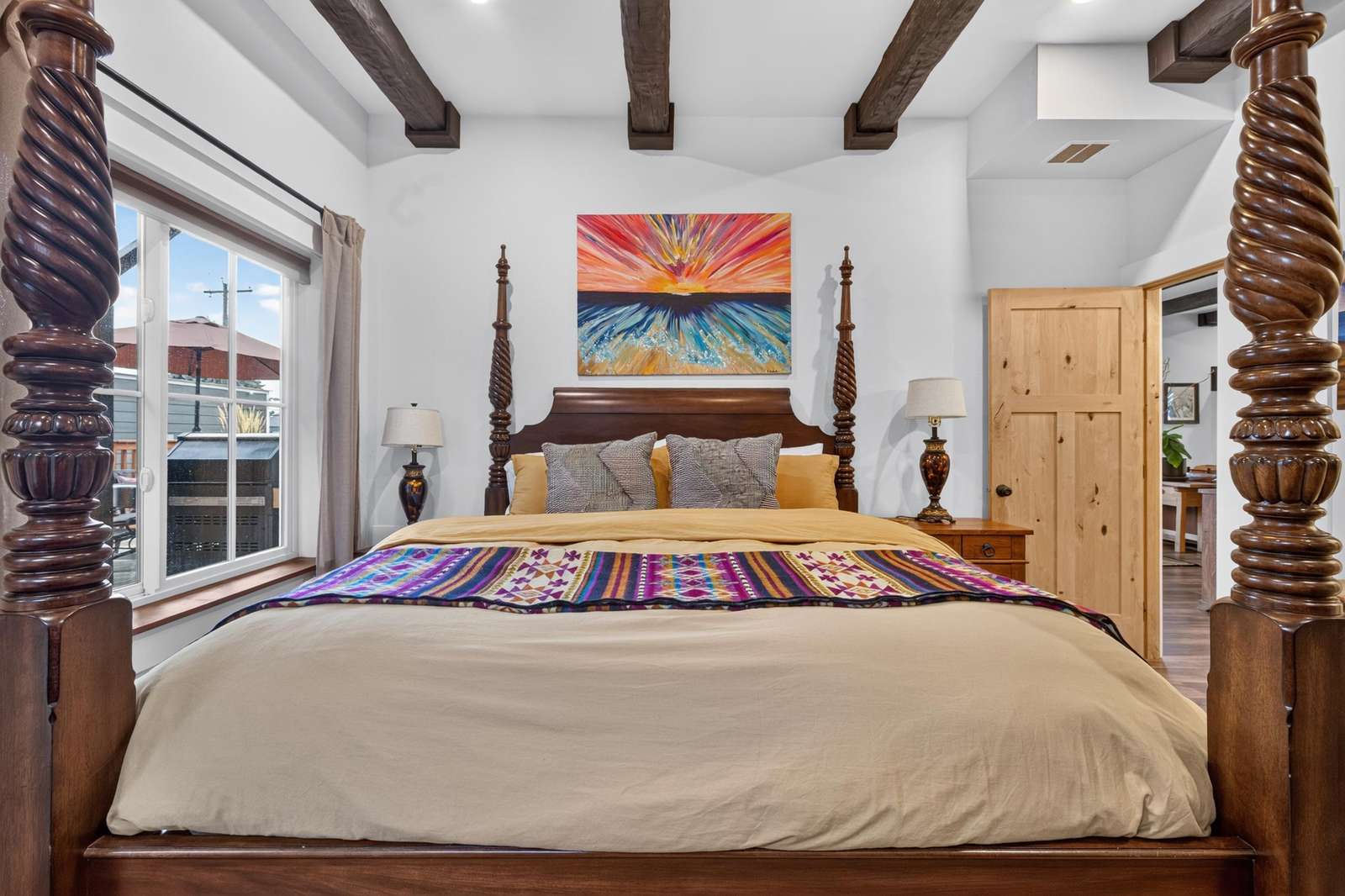 Dark wood four-poster bed with beige bedding and colorful throw, flanked by vintage lamps and sunset painting.