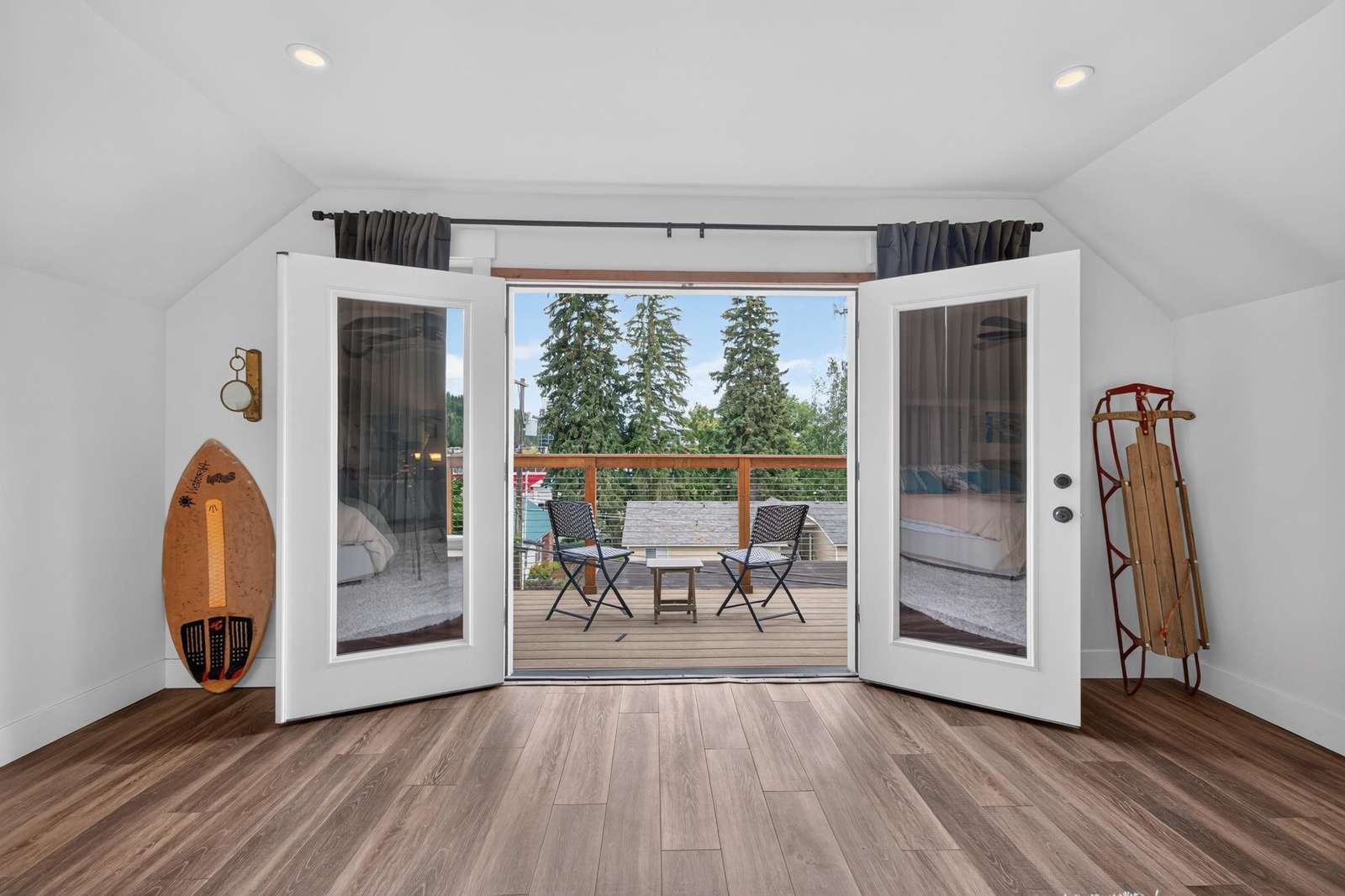 Loft area with wide plank floors, white walls, surfboard, vintage sled, and French doors opening to a wood balcony.