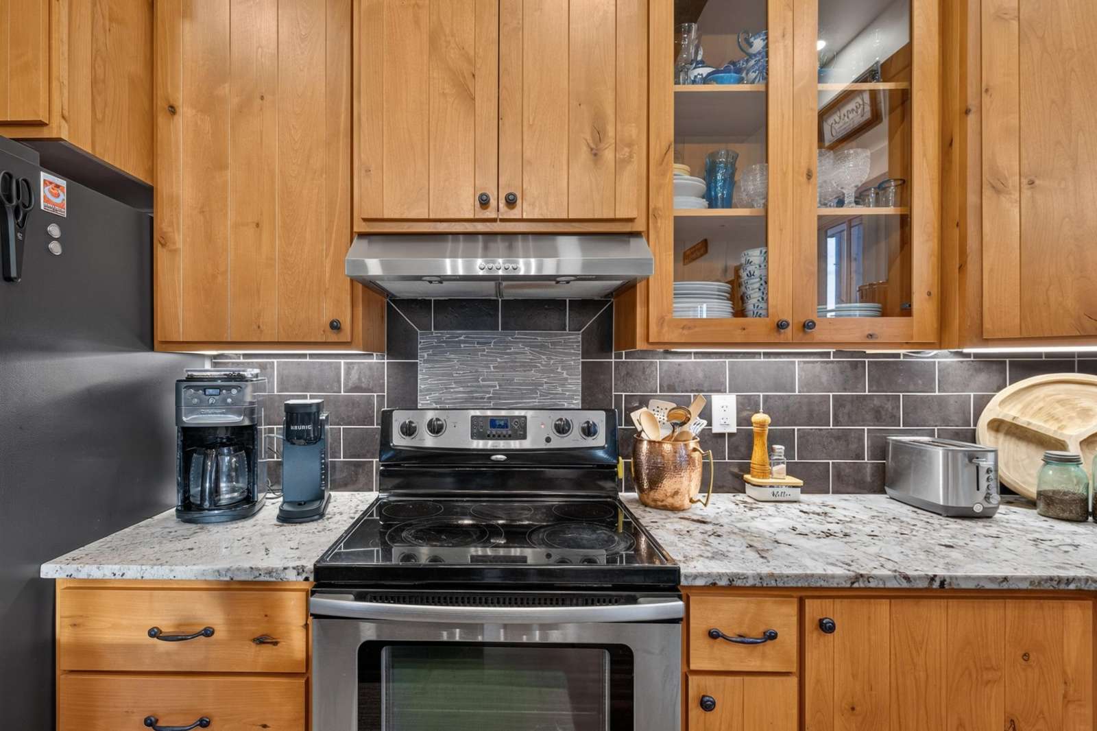 Electric stove with stainless steel range hood, granite countertops, and wood cabinetry with glass fronts.