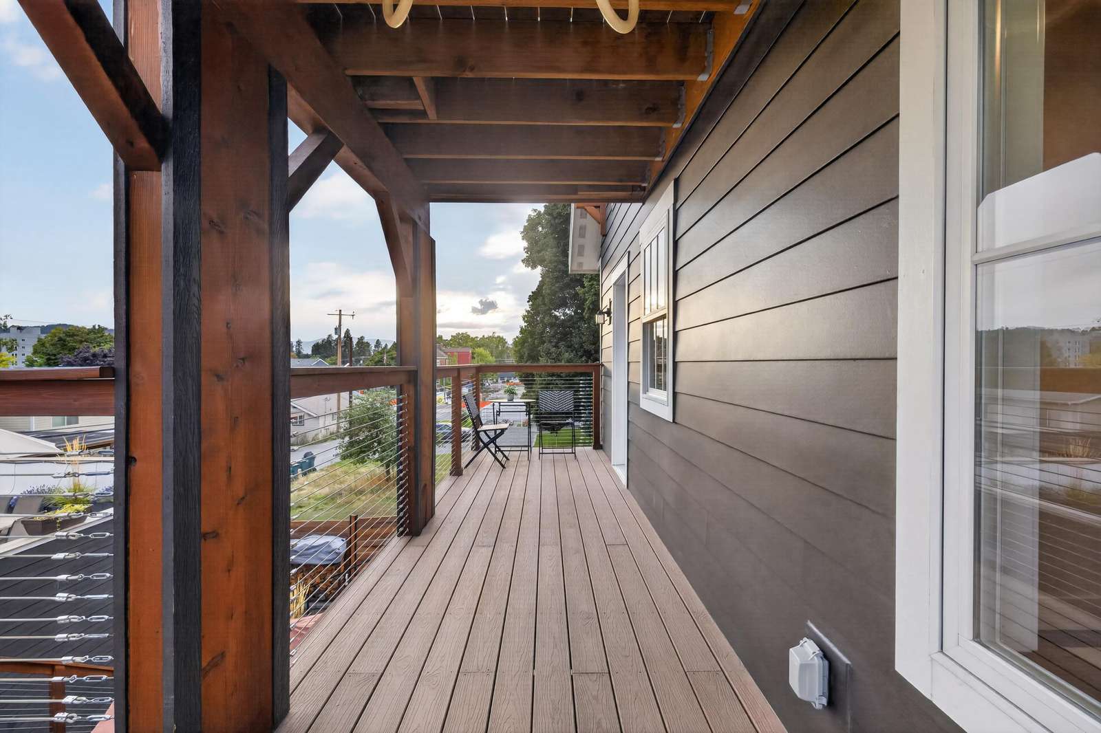 Balcony with wood plank flooring, cable railing, dark brown siding, and two black metal chairs at the far end.