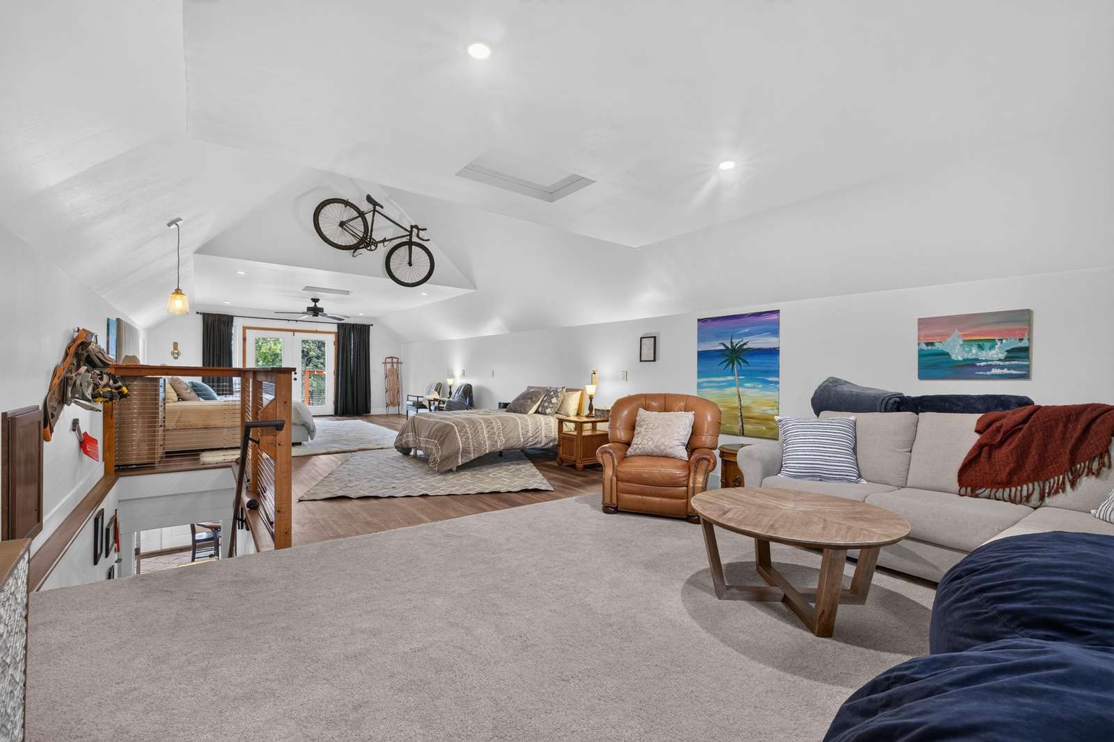 Loft space with vaulted ceilings, a mounted bike, beige sectional, leather armchair, and queen bed on wood floors.
