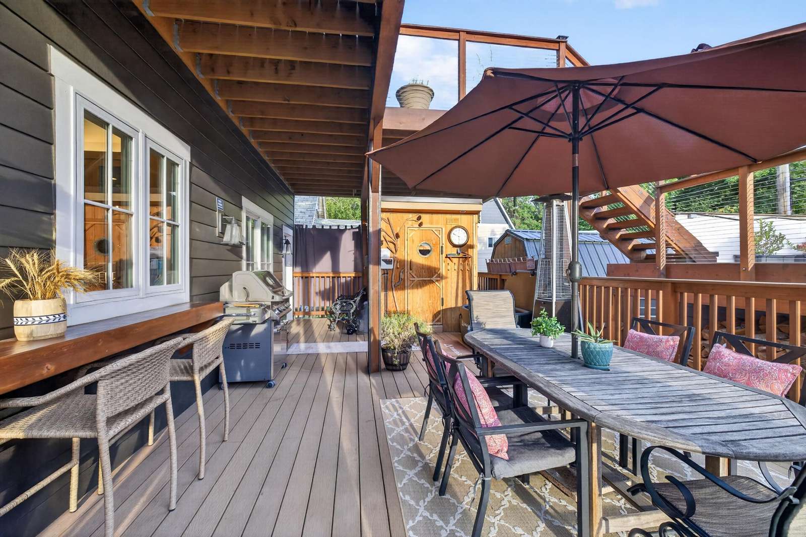 Outdoor deck with gray wood dining table, black chairs with pink cushions, large brown umbrella, and grill.