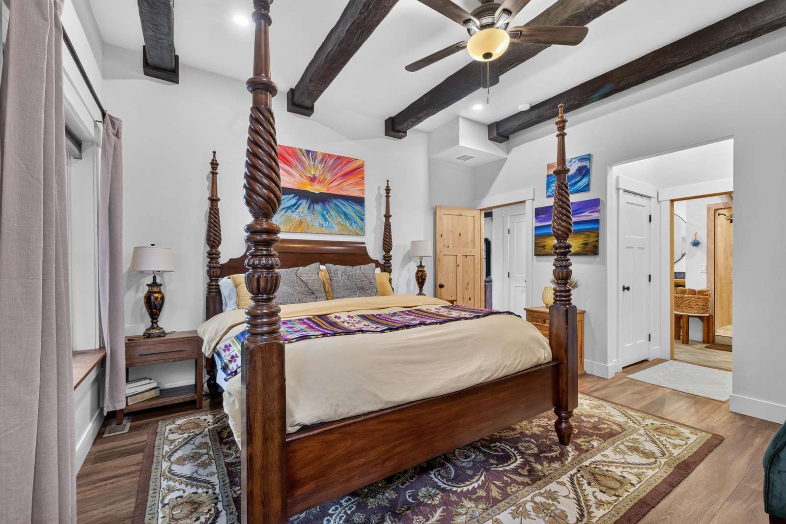 Four-poster wooden bed anchors the bedroom beneath dark ceiling beams, with colorful paintings and patterned rug.