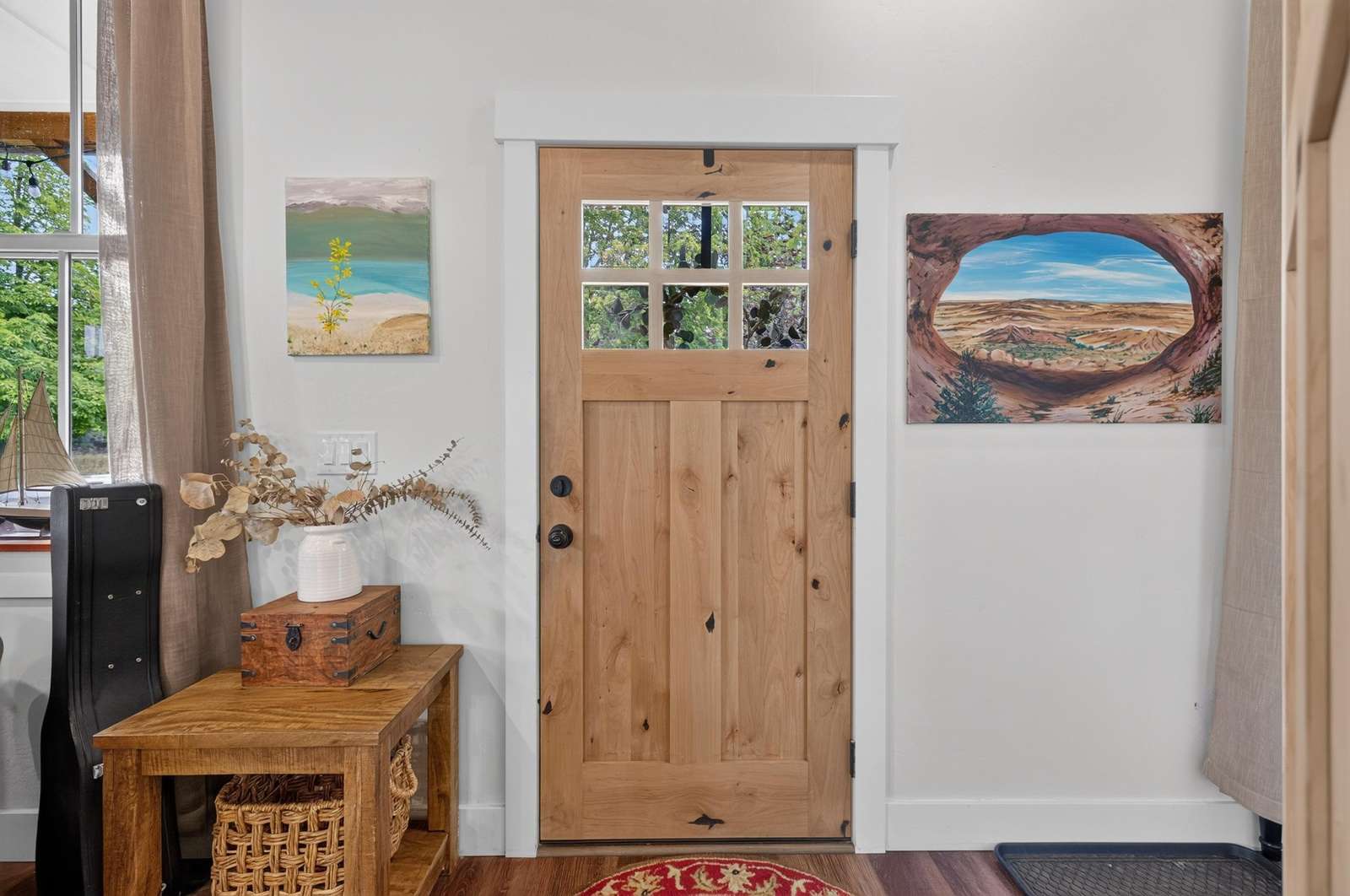 Front entryway with a wooden door featuring six glass panes, flanked by landscape paintings and a wicker basket console.
