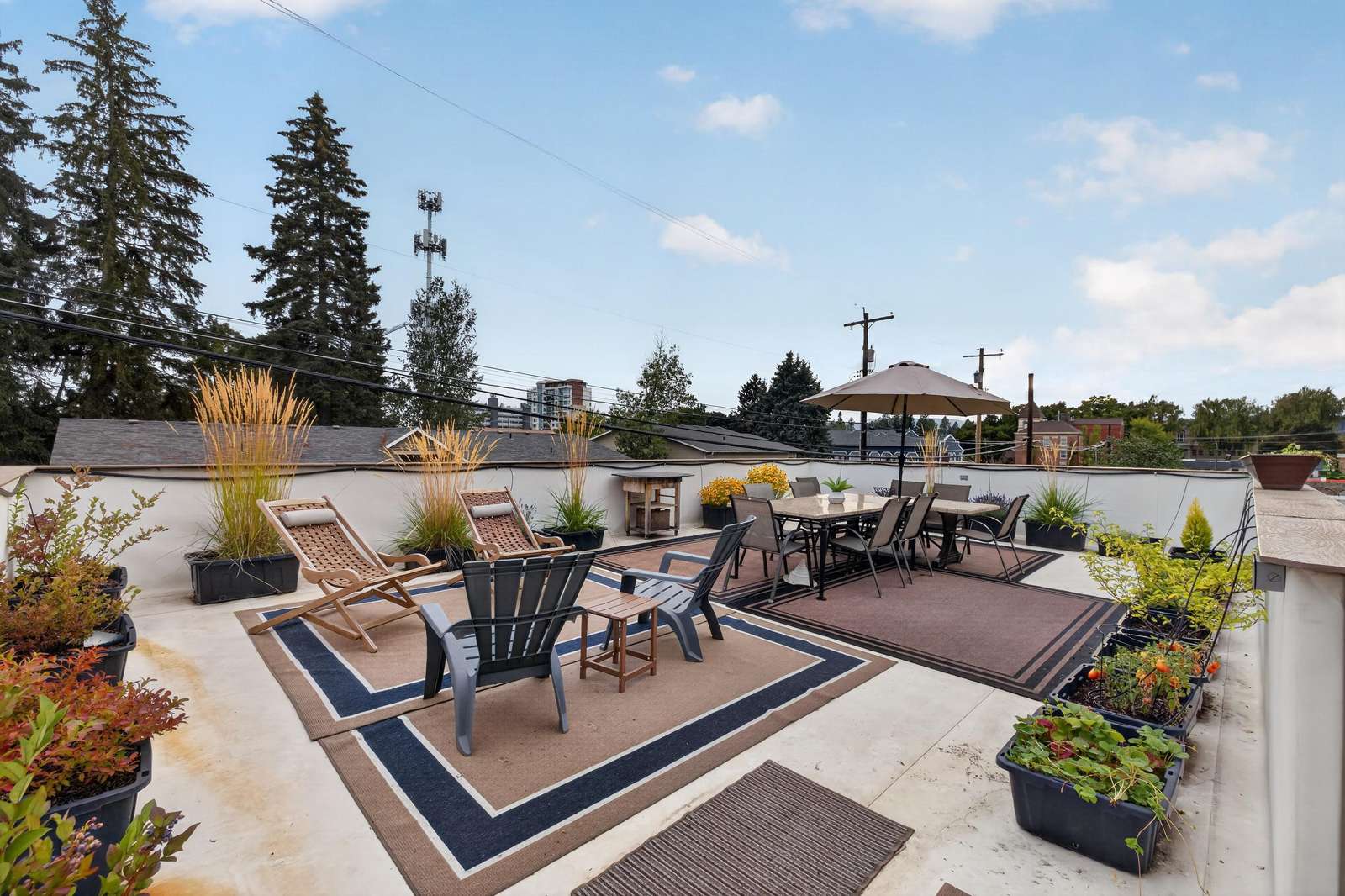 Rooftop patio with lounge chairs, rectangular dining table under umbrella, and potted plants along the perimeter.