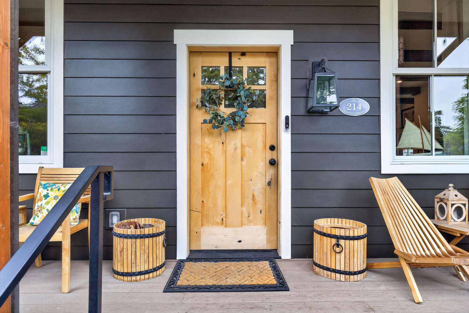 Natural wood front door with square windowpanes, flanked by black siding, wooden seating, and decorative barrels.
