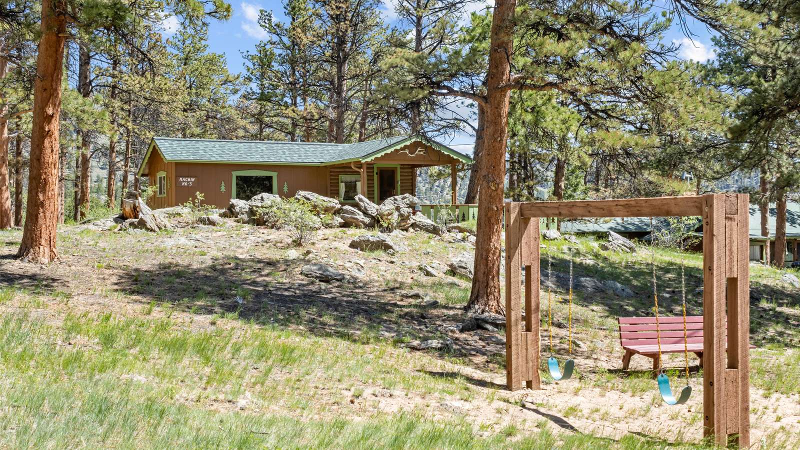 Outdoor play area with wooden swing set, red bench, and grassy yard framed by pine trees. - property