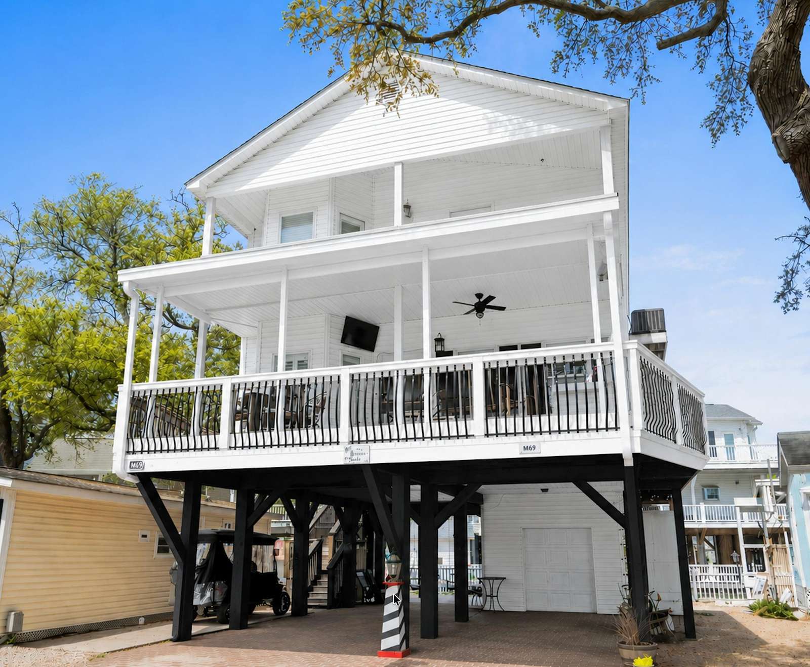 Two-story elevated white house with wraparound balcony, black metal railings, ceiling fan, and outdoor TV. - property