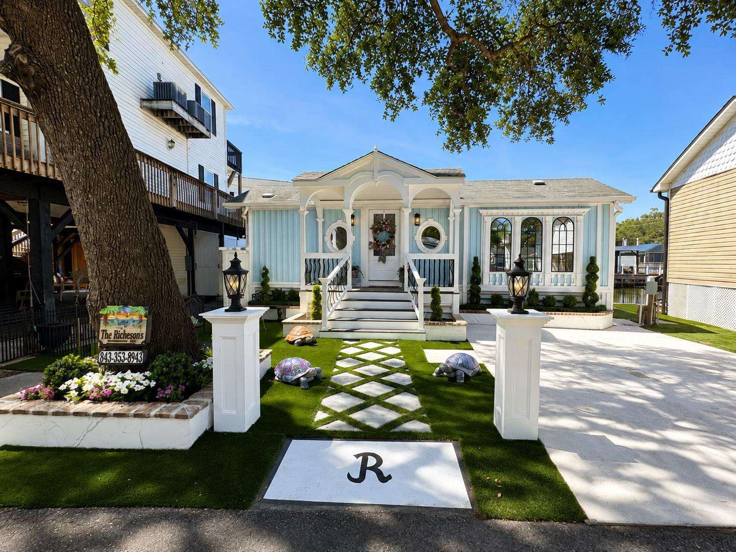 Pathway of diamond-patterned white tiles cuts through green artificial turf to the blue house’s front porch with circular windows. - property