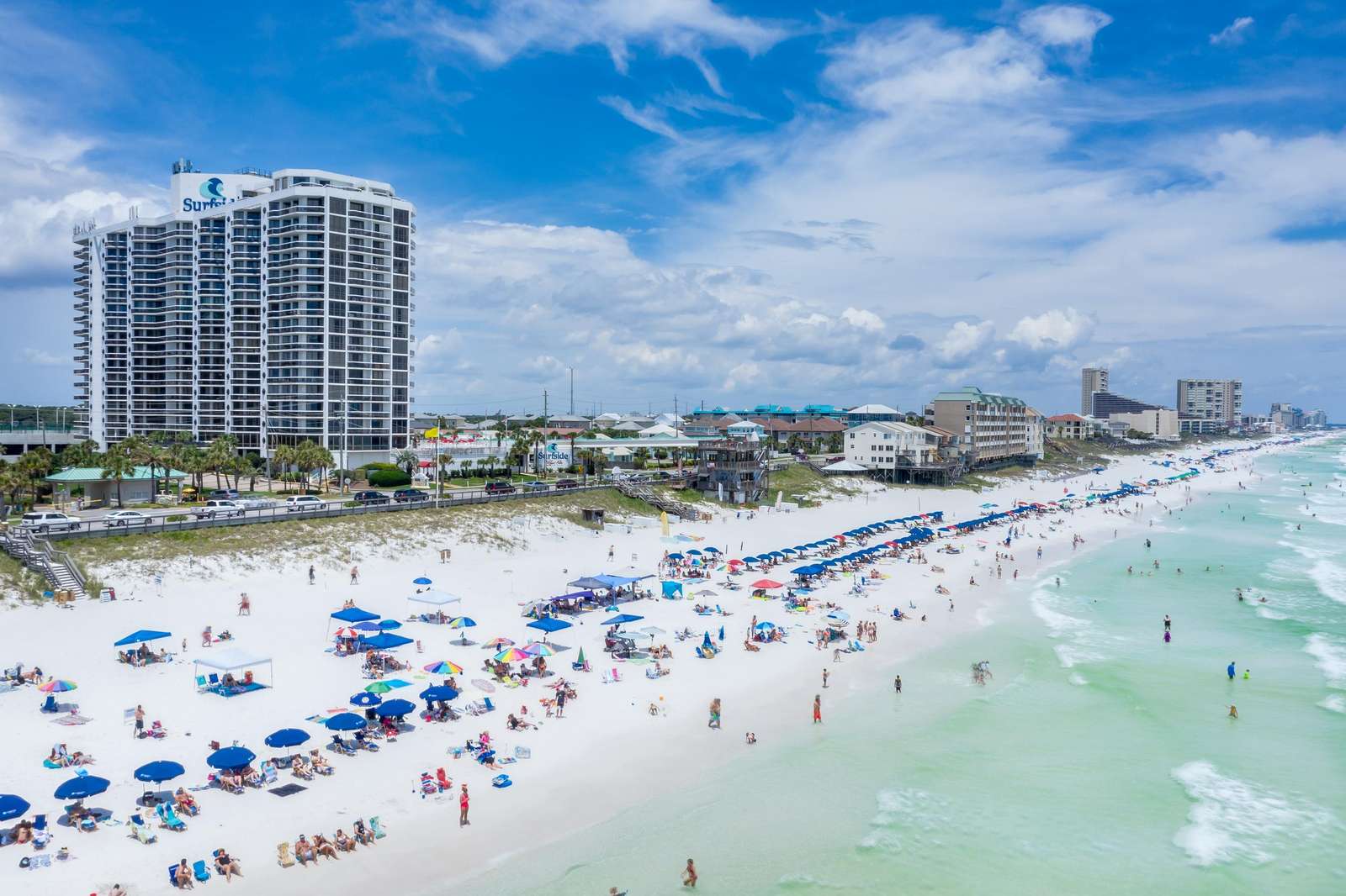 Wide view of Surfside Resort 1212 with white sandy beach, blue umbrellas, and turquoise Gulf waters in Miramar Beach. - property