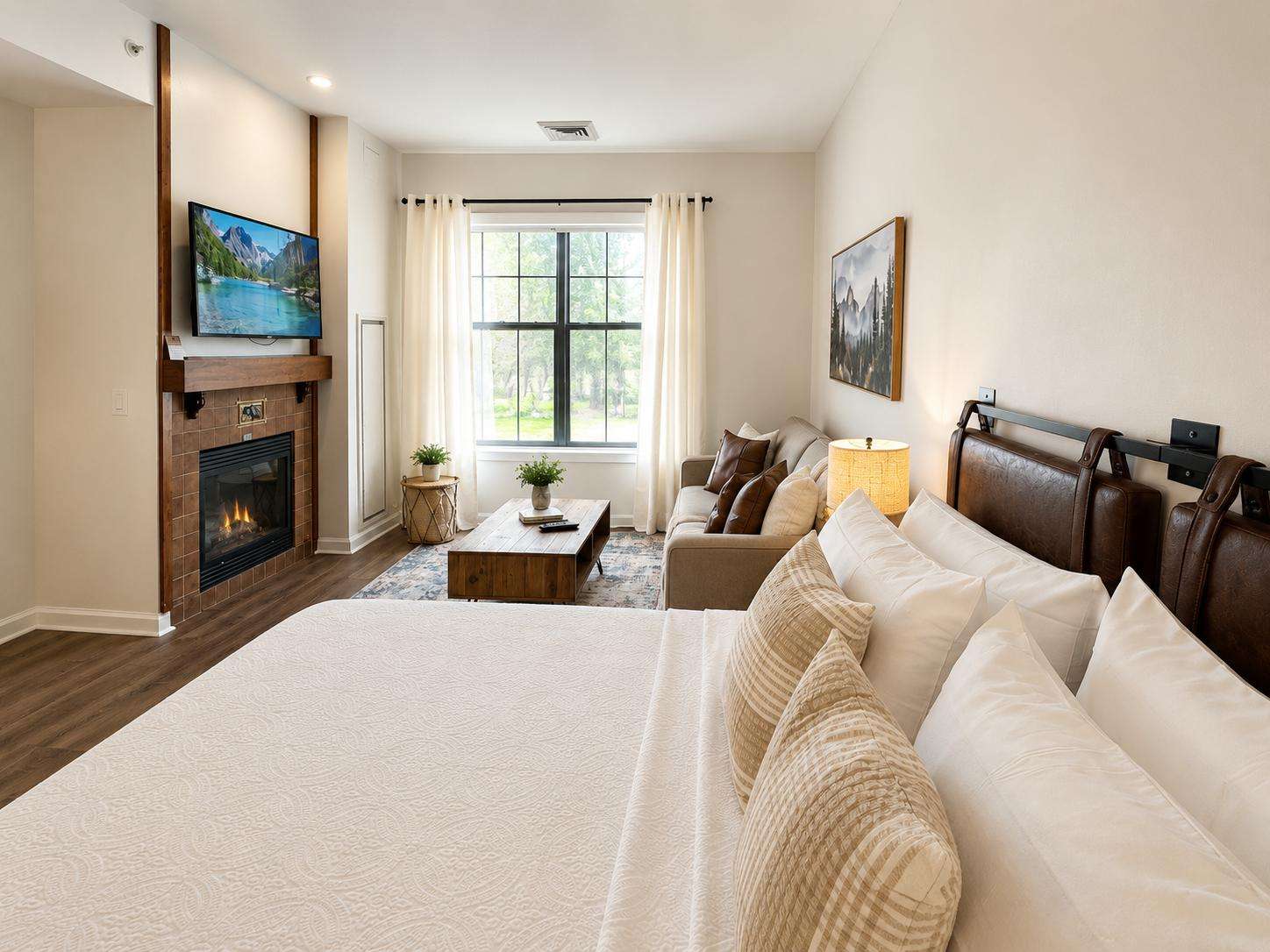 King bed with leather headboard faces a gas fireplace and wall-mounted TV, natural light from a large window. - property