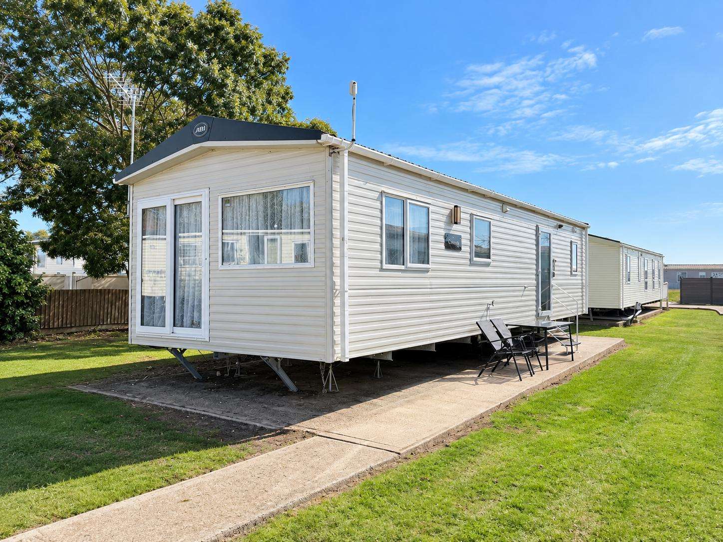 Exterior view of the white mobile home with large windows, patio chairs, and a paved walkway on green lawn. - property