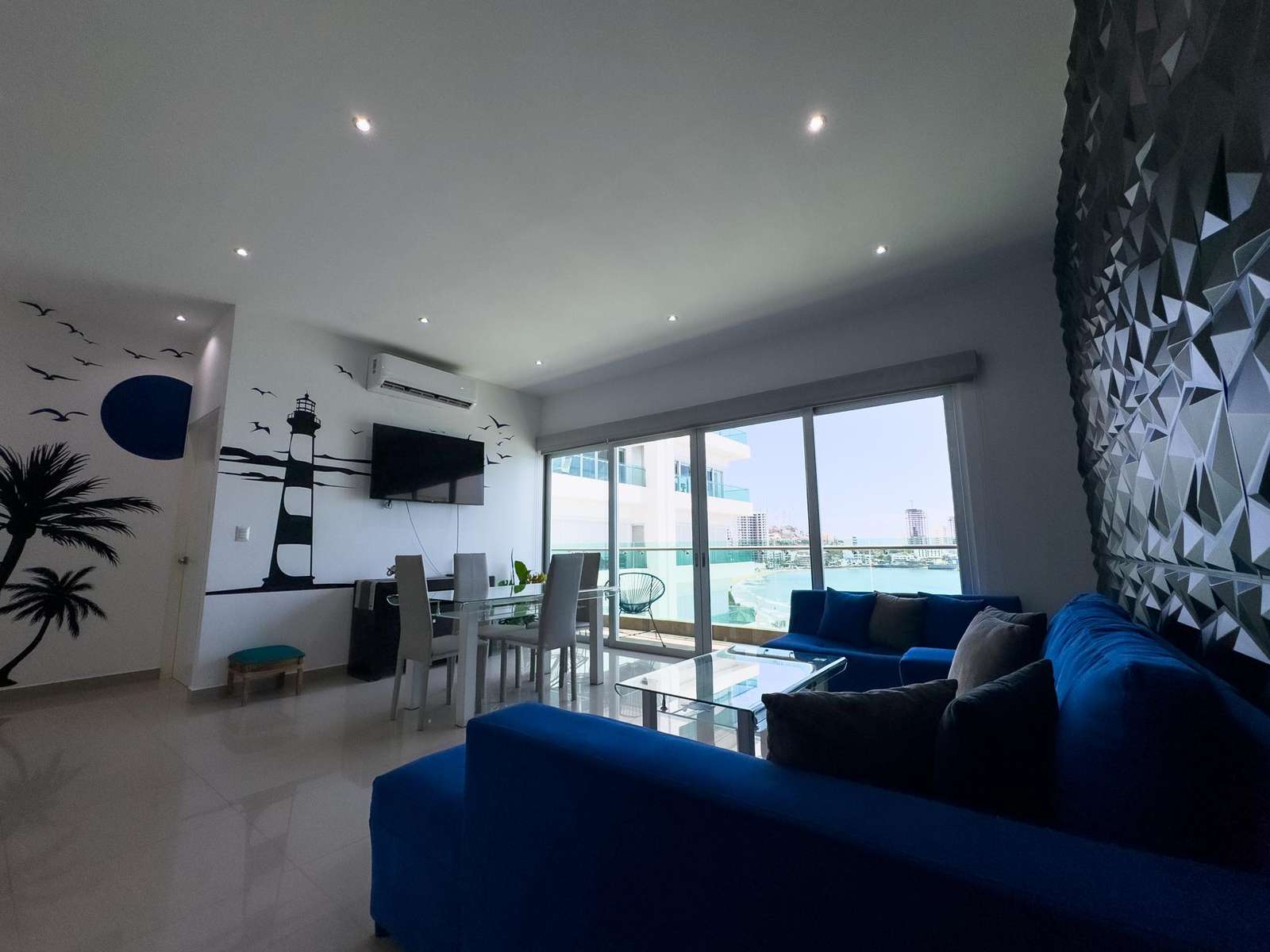 Living room features a deep blue sectional, geometric textured wall, glass dining table, and ocean views through sliding doors. - property
