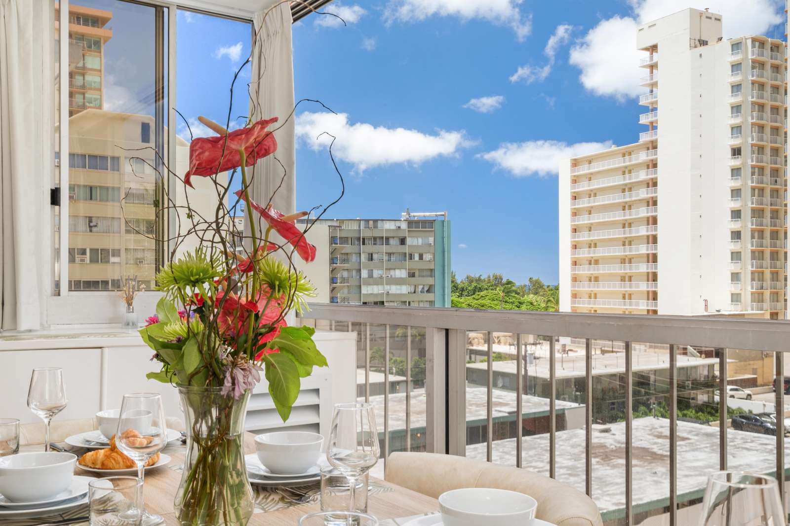 Dining table set with white bowls, glassware, and a vase of red and green tropical flowers near the balcony railing.