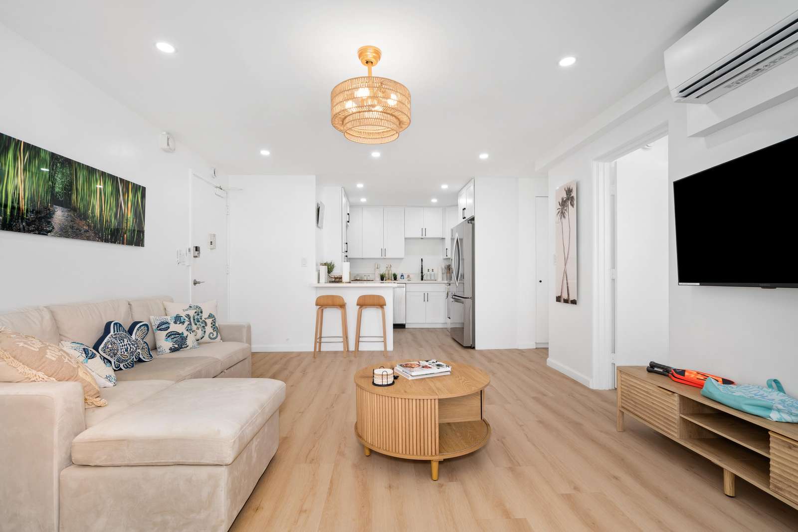 Living area with light wood flooring, beige sectional sofa, round wood coffee table, and wall-mounted TV under A/C unit.