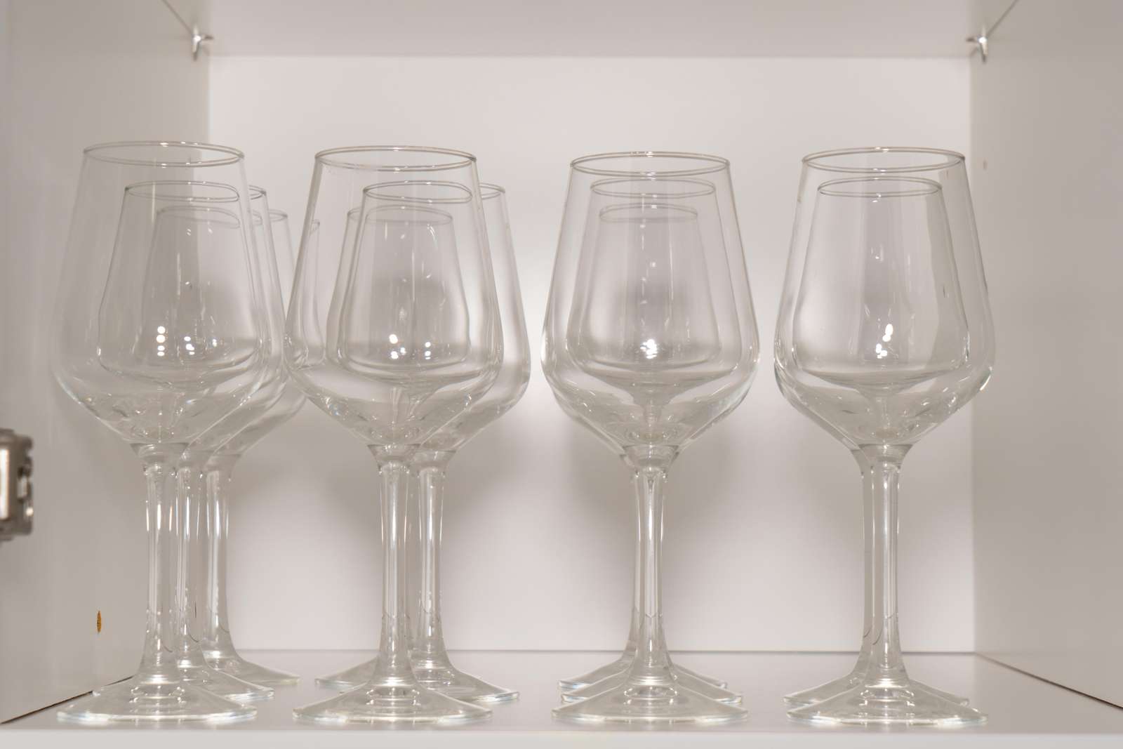 Clear wine glasses neatly arranged on a white kitchen cabinet shelf with soft overhead lighting.