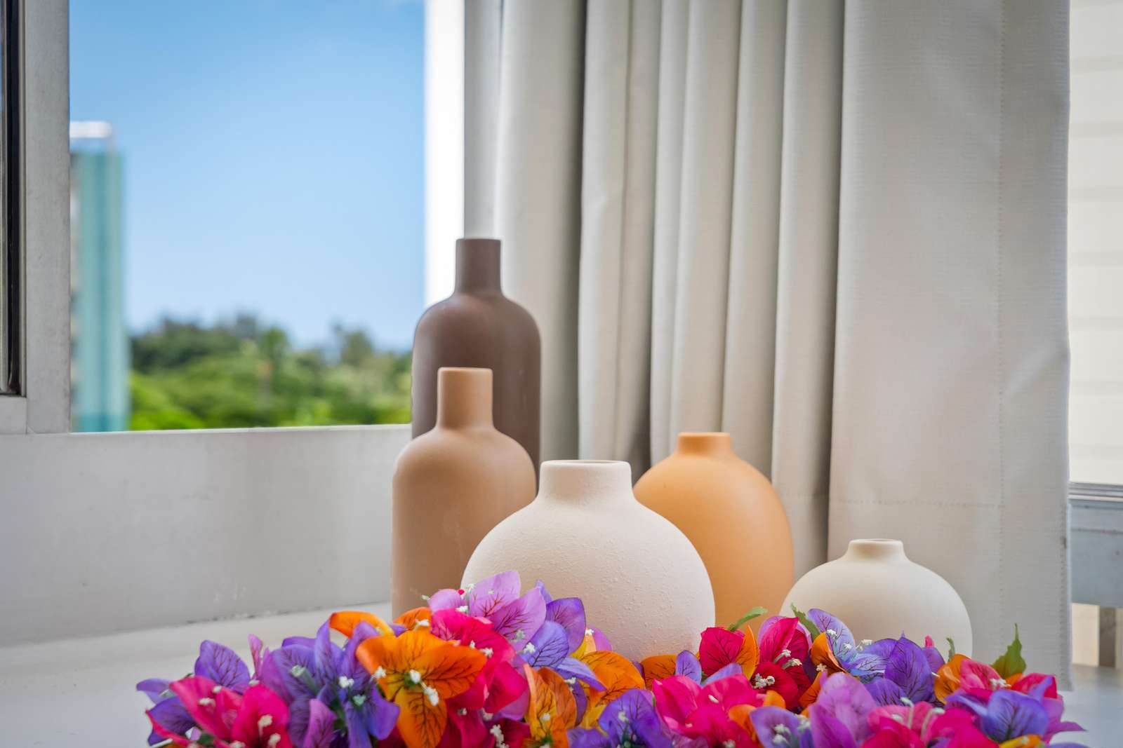 Earth-toned ceramic vases cluster on a white ledge beside vibrant purple, orange, and pink flowers near a window.