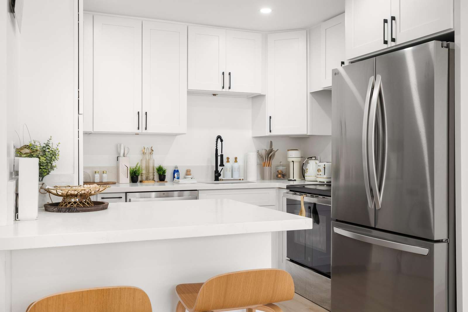 Quartz countertop peninsula extends with two light wood stools beneath white shaker cabinets and stainless steel appliances.