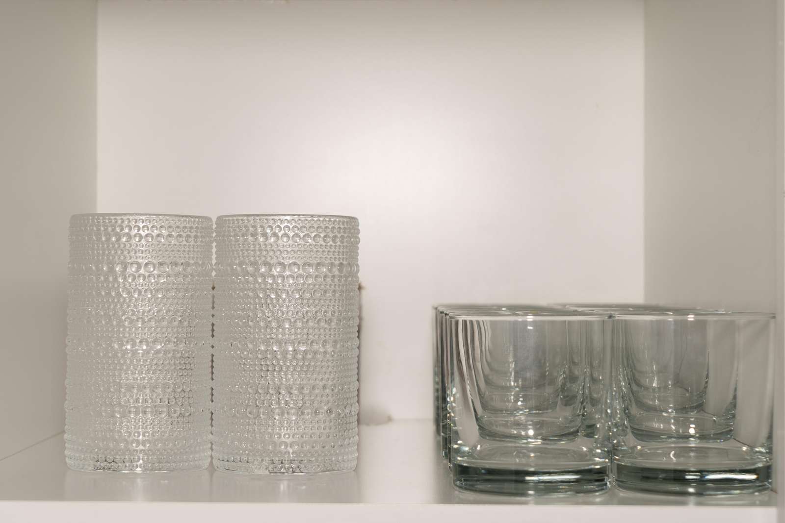 Textured glass tumblers stand beside smooth clear square glasses on a white cabinet shelf.