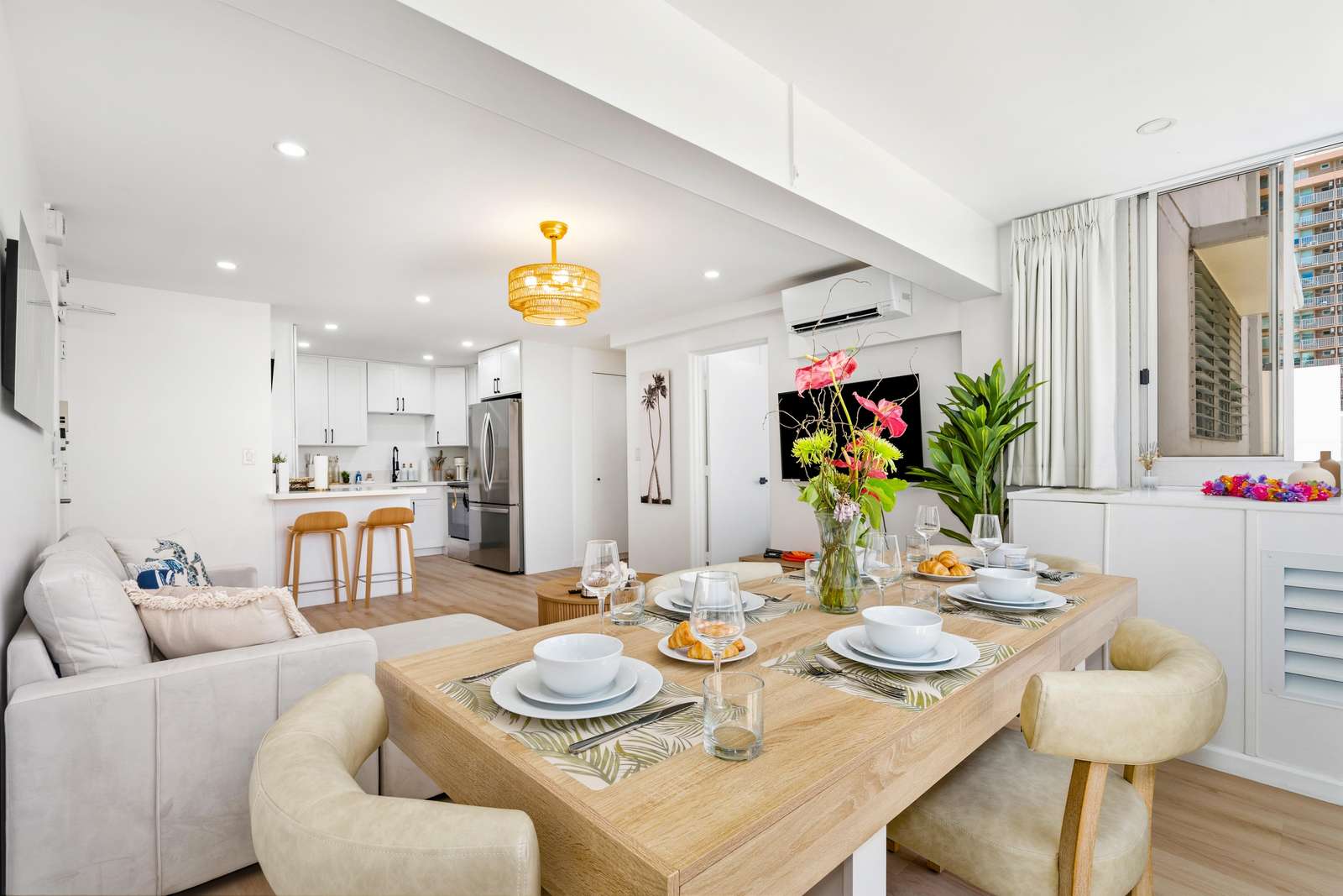 Natural light fills the open living and dining area with cream leather chairs, light wood floors, and tropical floral centerpiece.