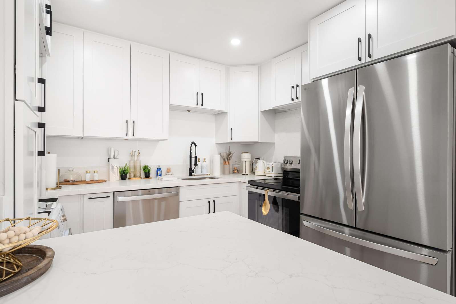 White quartz island faces stainless steel French-door refrigerator and black faucet sink beneath white shaker cabinets.