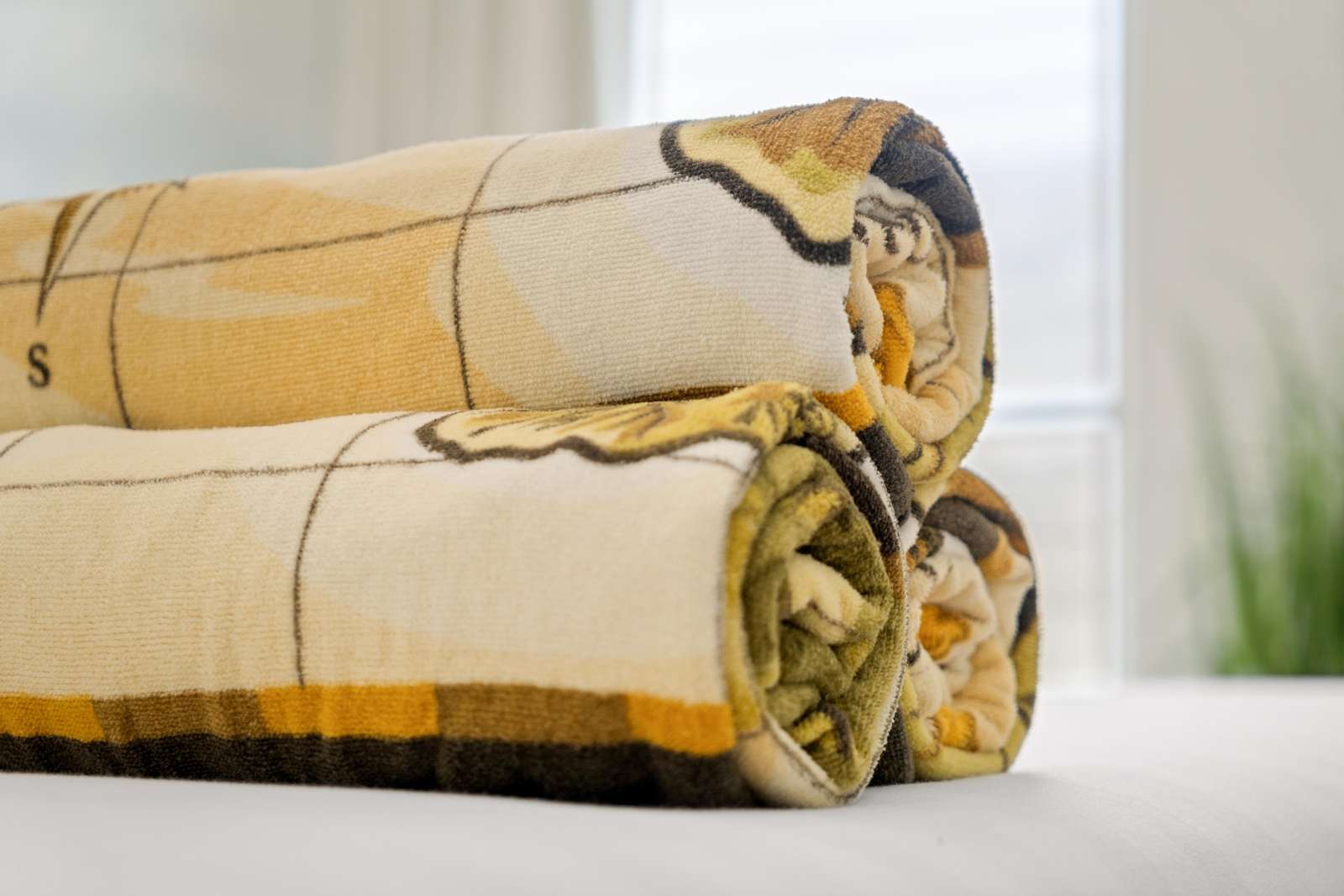 Three rolled towels with yellow and brown patterns sit on a white bedspread near a bright window.
