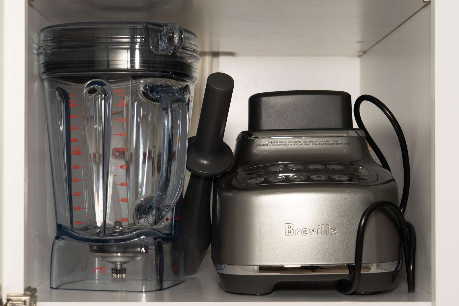 Silver Breville blender base and clear blender pitcher with black tamper stored inside white kitchen cabinet.