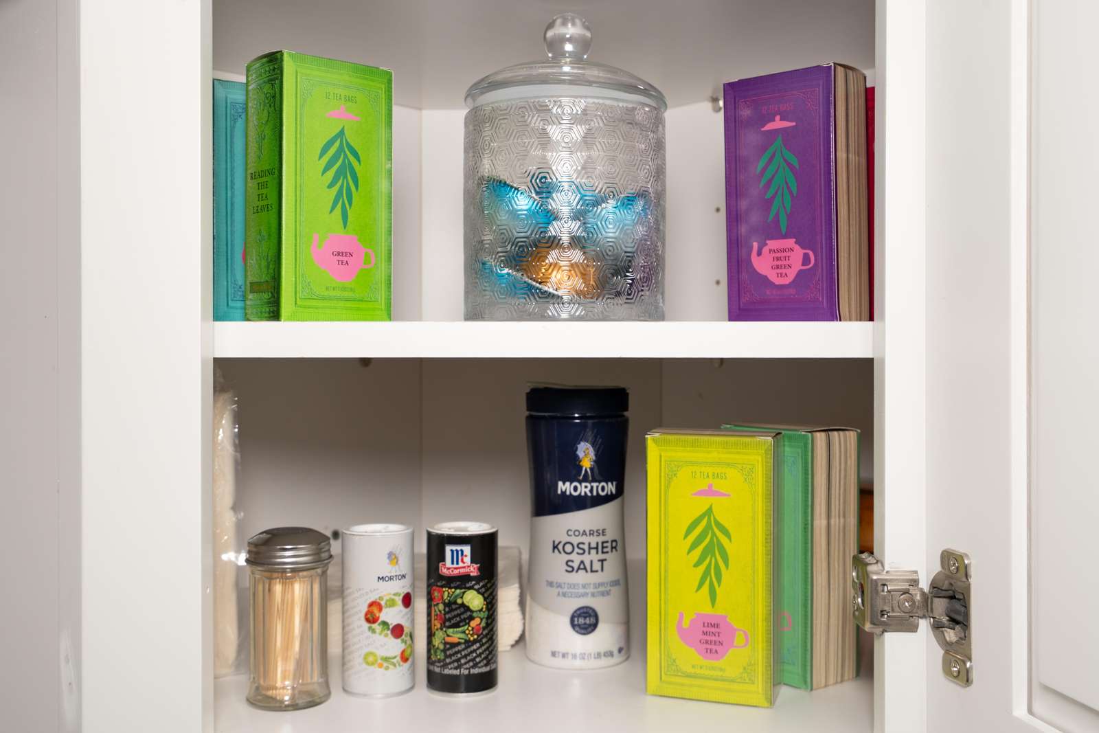 White kitchen cabinet shelves hold colorful tea boxes, a textured glass jar, and assorted spices and salt.