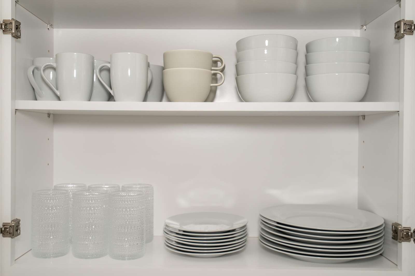 White kitchen cabinet shelves hold stacked white mugs, bowls, textured glass tumblers, and plates in organized rows.