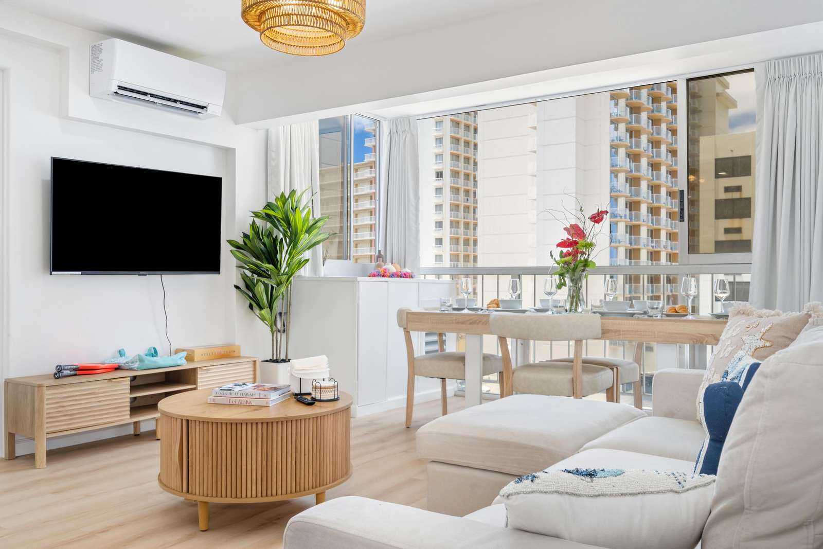 Sliding floor-to-ceiling windows reveal city views beyond the light wood dining table set with glassware and a floral centerpiece.