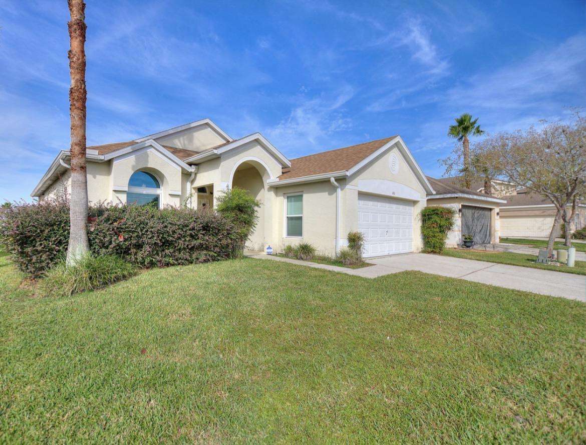 Single-story light beige house with arched entryway, two-car garage, and manicured front lawn under blue sky. - property