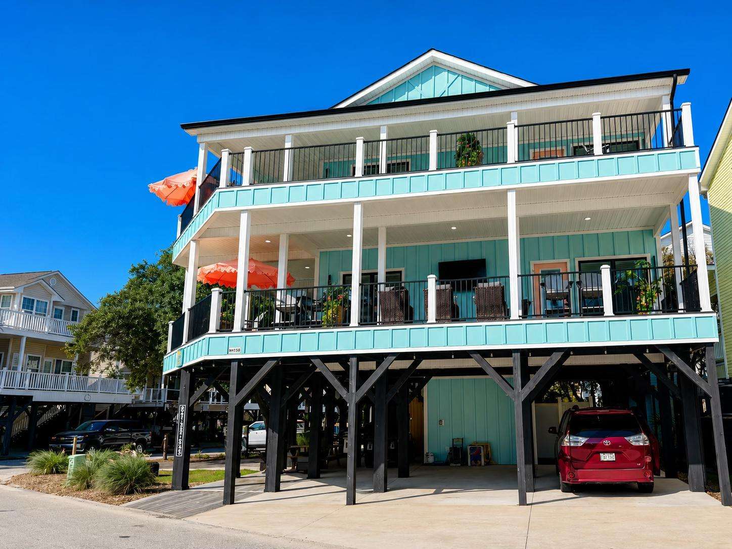 Two-story elevated beach house with pale turquoise siding, black stilts, wraparound balconies, and coral scalloped umbrellas. - property